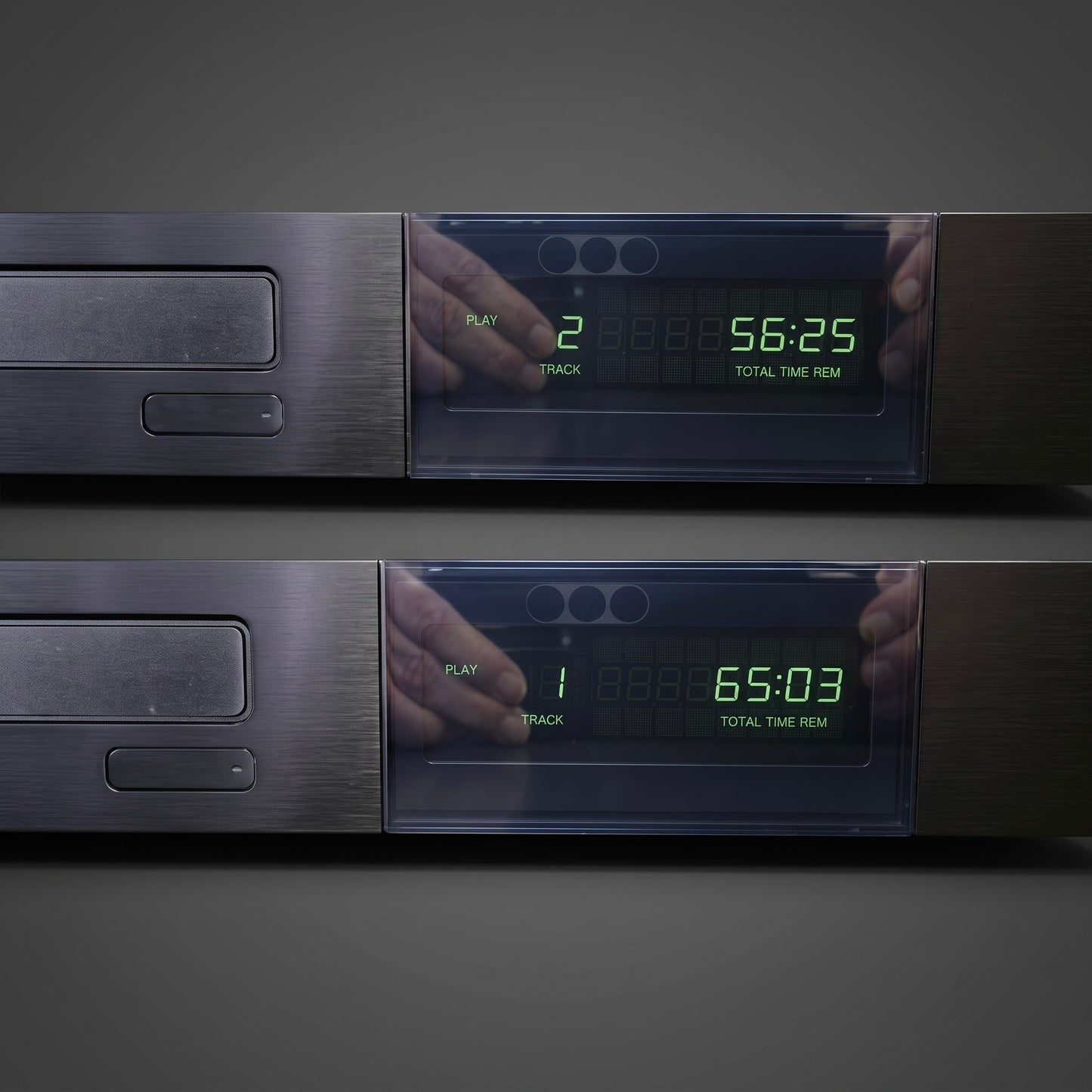 Two dark gray digital audio players or CD players shown stacked vertically against a dark gray background. Each unit has a brushed metal front, a transparent display window, and green LED numbers showing track and time information.