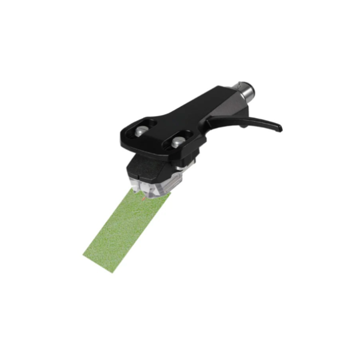 A high-angle studio shot against a plain white background shows a black phono cartridge mounted on a headshell, with its needle resting on a small, rectangular strip of light green abrasive paper, known as "Linn Green Paper." The cartridge is angled toward the lower left, and the fine green paper is positioned diagonally beneath the stylus tip to demonstrate a specialized cleaning method for turntable needles.