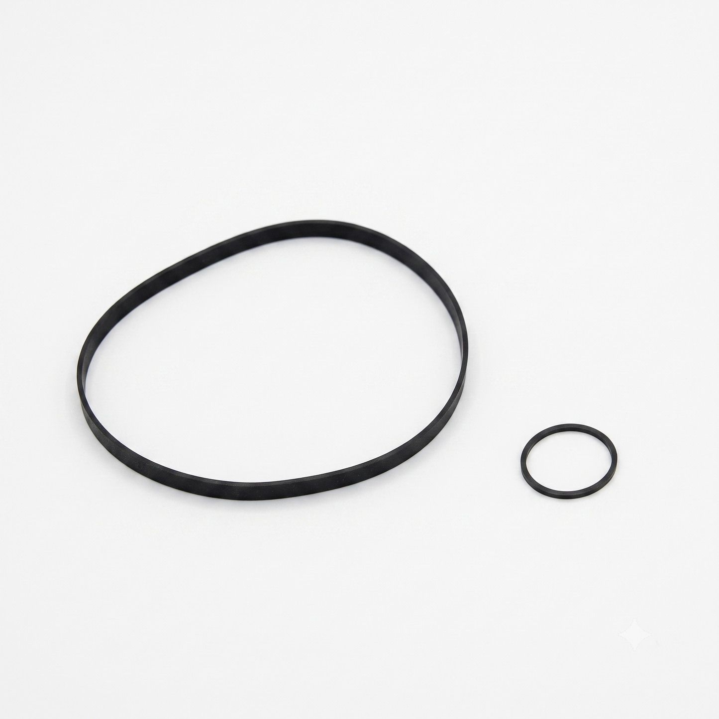 A black-and-white photograph shows a pair of black rubber belts from a Yamaha KX-650/KX-930 belt kit, isolated against a plain white background.
On the left is a large, thin, flat rubber drive belt forming a loose, asymmetrical oval shape. On the right sits a much smaller, thick, circular rubber belt or O-ring. Both items are viewed from a high angle, casting subtle shadows onto the surface beneath them.