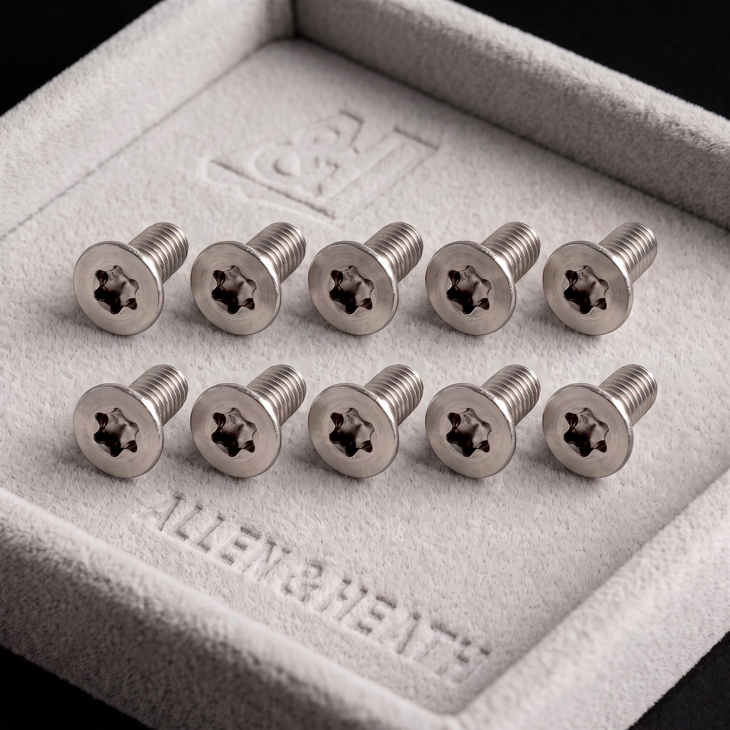 A close-up photograph of ten stainless steel, Allen & Heath fader screws AB3819 10-Pack with Torx (star) drive heads, neatly arranged in two rows of five on a plush grey velvet display tray.