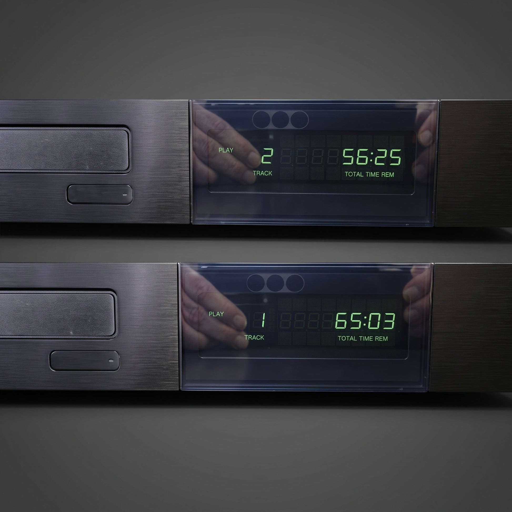Two dark gray digital audio players or CD players shown stacked vertically against a dark gray background. Each unit has a brushed metal front, a transparent display window, and green LED numbers showing track and time information.