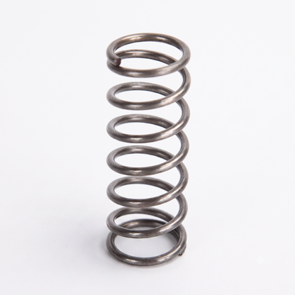 A dark metallic, helical coil spring stands vertically against a plain white background. The spring has approximately six distinct coils, with open spaces between them, and flat-ground ends at the top and bottom.