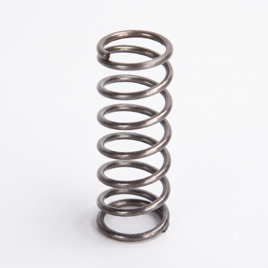 A dark metallic, helical coil spring stands vertically against a plain white background. The spring has approximately six distinct coils, with open spaces between them, and flat-ground ends at the top and bottom.
