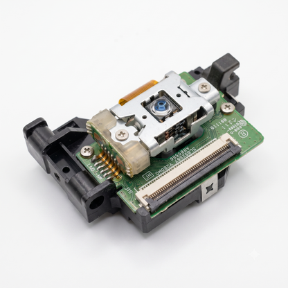 High-definition product photograph of an electronic optical disc drive component, likely a laser lens mechanism, isolated on a seamless white background. The device features a black plastic housing, a green printed circuit board (PCB) with a ribbon cable connector slot, and a central metal-framed laser assembly housing a clear, blue-tinted optical lens. Delicate solder joints, screws, and circuit markings are sharply visible.