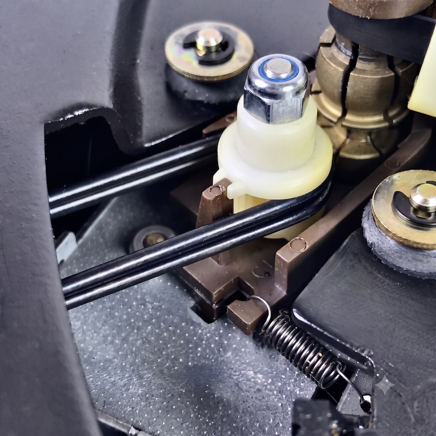 Close-up of a mechanical assembly with black rods, a cream plastic component, brass hardware, and a spring on a dark textured surface.