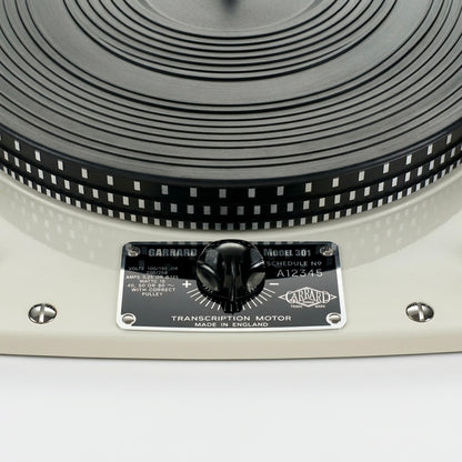 High-resolution close-up shot of a restored vintage Garrard Model 301 Schedule No 9 transcription motor turntable. The image features the iconic glossy cream-white chassis, a textured black rubber platter mat with a strobe rim pattern, and a highly polished black and silver faceplate. A central black pitch control knob sits on the faceplate alongside technical specifications and the classic Garrard logo. Two polished chrome slotted mounting bolts are visibly flush on the left and right sides of the chassis.