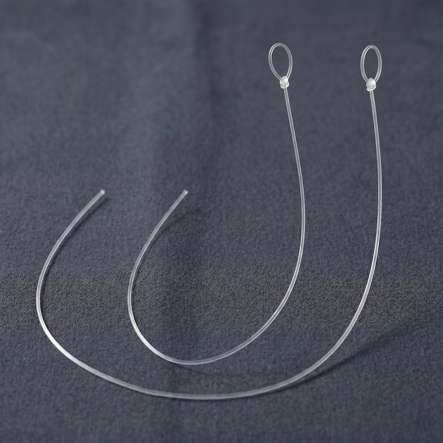 A close-up photograph featuring two identical, replacement anti-skate threads for a turntable, presented as a 2-pack. Both translucent, ultra-thin monofilament nylon lines are arranged side-by-side in parallel, semi-circular curves on a dark grey, textured fabric background. Each line is identically sized, cleanly cut at one open end on the left, and features a pre-tied loop secured with a small, precise knot at the top right.