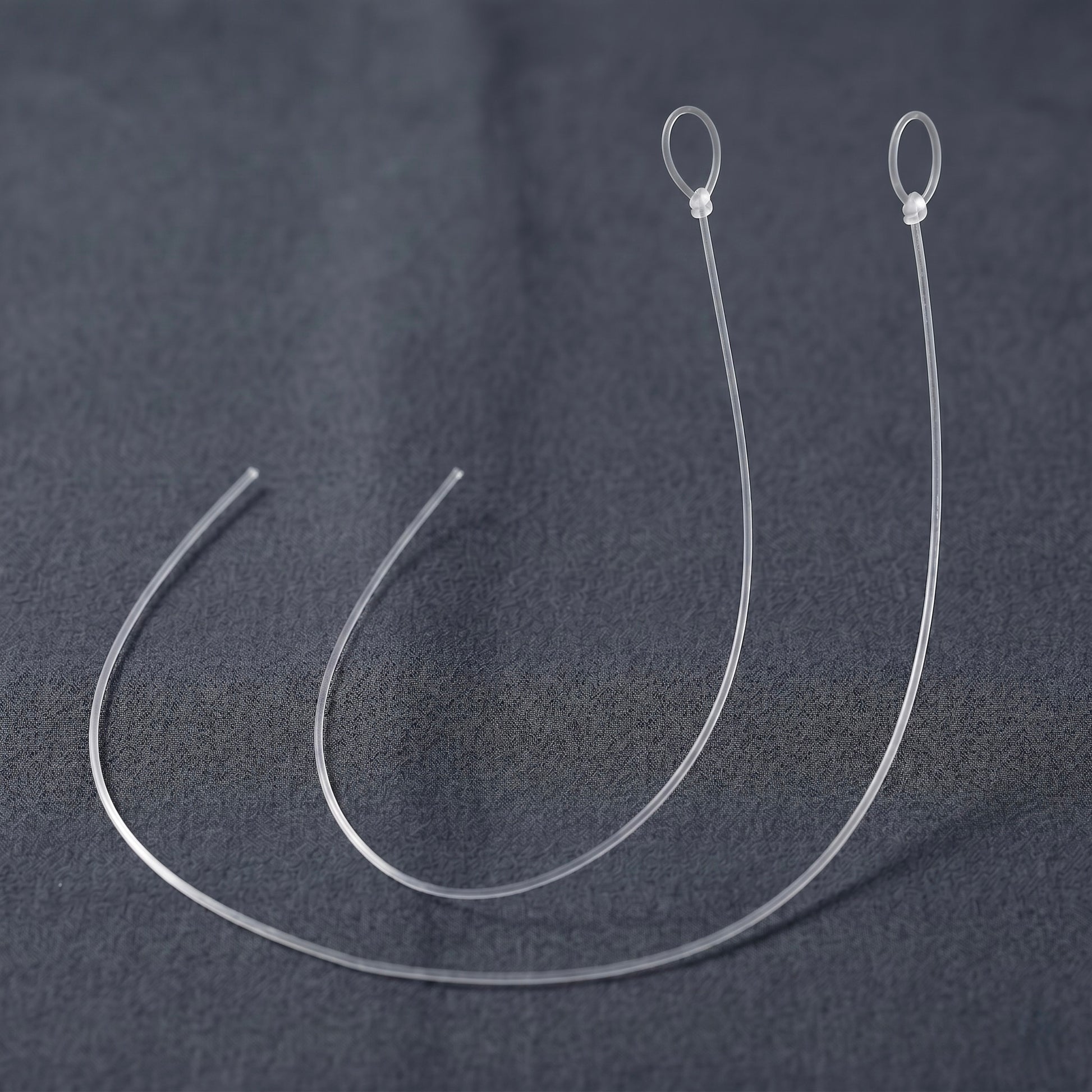 A close-up photograph featuring two identical, replacement anti-skate threads for a turntable, presented as a 2-pack. Both translucent, ultra-thin monofilament nylon lines are arranged side-by-side in parallel, semi-circular curves on a dark grey, textured fabric background. Each line is identically sized, cleanly cut at one open end on the left, and features a pre-tied loop secured with a small, precise knot at the top right.