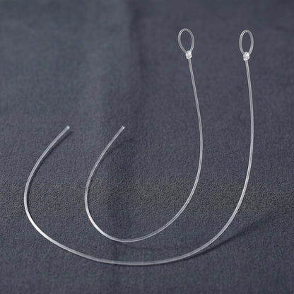 A close-up photograph featuring two identical, replacement anti-skate threads for a turntable, presented as a 2-pack. Both translucent, ultra-thin monofilament nylon lines are arranged side-by-side in parallel, semi-circular curves on a dark grey, textured fabric background. Each line is identically sized, cleanly cut at one open end on the left, and features a pre-tied loop secured with a small, precise knot at the top right.