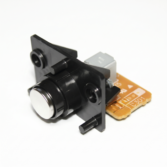 A close-up studio shot against a seamless white background showing the front side of a Technics reset button assembly. The unit features a prominent, circular silver/metallic button housed within a black plastic mounting bracket. Two small plastic alignment pins and two screw holes are visible on the bracket's face. Peeking out from behind the black plastic assembly is a small, light-brown printed circuit board (PCB).