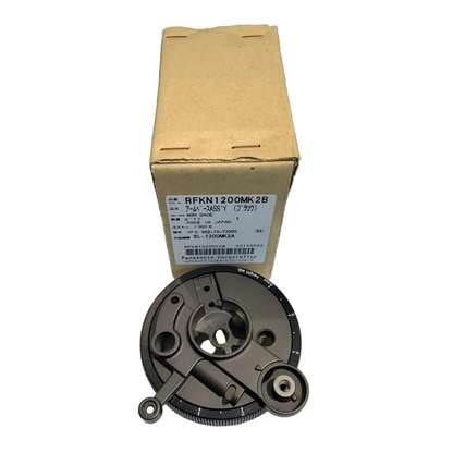 A top-down view of a Technics RFKN1200MK2B tonearm base resting in front of its original cardboard packaging. The black, circular mechanical part shows its complex mounting structure and height adjustment dial with white numerical markings. The vertical brown box behind it features a white manufacturer label with the part number "RFKN1200MK2B," the description "ARM BASE," and text indicating it is "MADE IN JAPAN" by Panasonic Corporation.