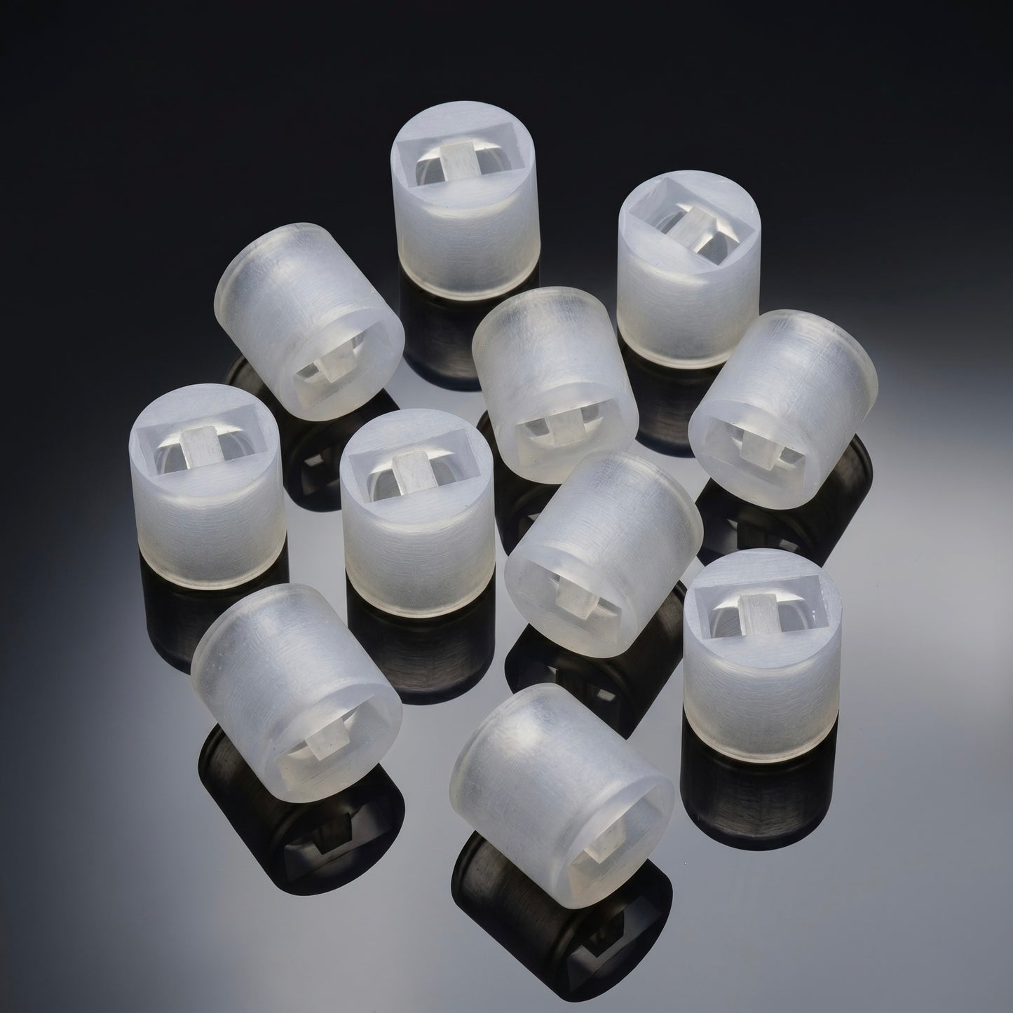 close-up studio shot of ten identical, small cylindrical spacers arranged together on a glossy, reflective dark grey surface. The spacers are made of a translucent, frosted white plastic or silicone material. Each piece features a hollow interior with a rectangular cutout and a small central dividing tab. Clear, dark reflections of each cylinder are cast directly beneath them on the shiny surface.