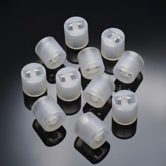 close-up studio shot of ten identical, small cylindrical spacers arranged together on a glossy, reflective dark grey surface. The spacers are made of a translucent, frosted white plastic or silicone material. Each piece features a hollow interior with a rectangular cutout and a small central dividing tab. Clear, dark reflections of each cylinder are cast directly beneath them on the shiny surface.