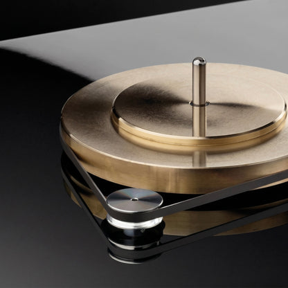 A close-up, high-angle shot of a premium turntable mechanism. It features a heavy, tiered sub-platter made of brushed brass with a polished silver spindle rising from the center. A flat black rubber drive belt is wrapped around the lower edge of the brass platter and loops around a smaller, polished silver motor pulley in the foreground. The entire assembly sits on a high-gloss, reflective black turntable base that perfectly mirrors the components.