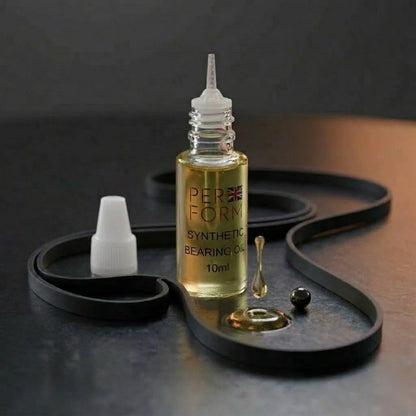 
A small dropper bottle of "PERFORM Synthetic Bearing Oil" (10ml) sits upright on a dark surface. The bottle contains a clear golden liquid and features a British flag logo. Surrounding the bottle is a black rubber drive belt arranged in a curved shape. In the foreground, a single drop of oil is suspended above a small puddle, next to a small black bearing ball. A white screw-cap for the bottle sits to the left.