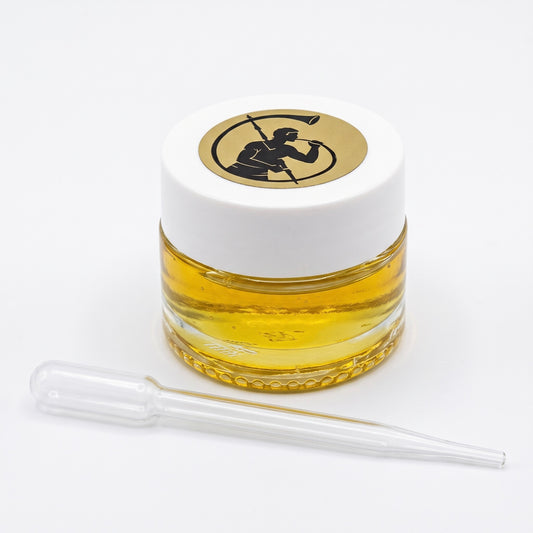 A small, clear glass jar filled with golden yellow liquid, topped with a white lid featuring a circular black-and-gold logo, with a clear glass pipette placed in front on a white background.