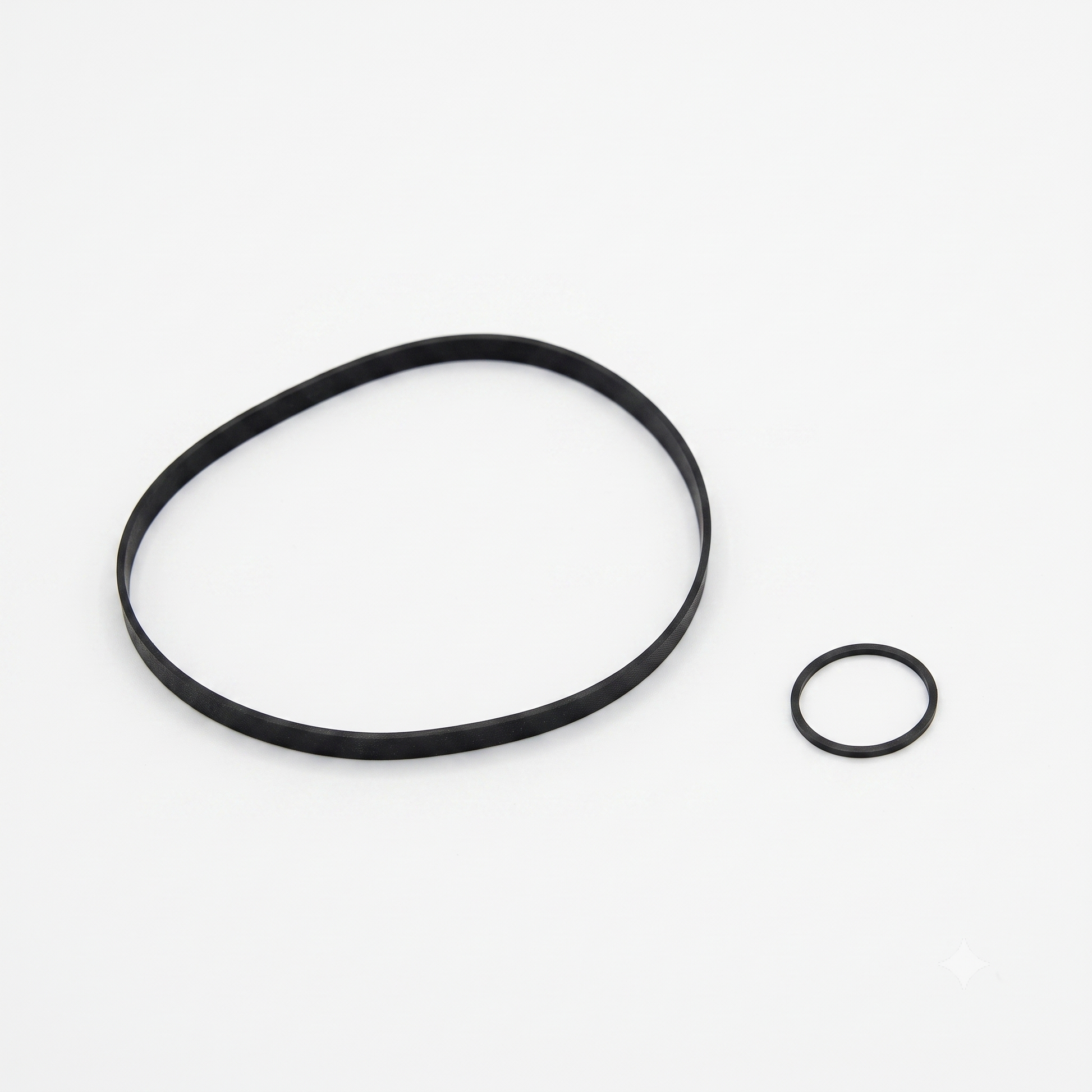 top-down, studio shot on a solid white background showing two black rubber replacement belts for a cassette deck.

On the left is a large, thin, flat capstan belt forming a loose, irregular oval shape. On the right sits a much smaller, square-cut rubber counter or assist motor belt forming a perfect circle. Both belts are matte black with a clean texture.